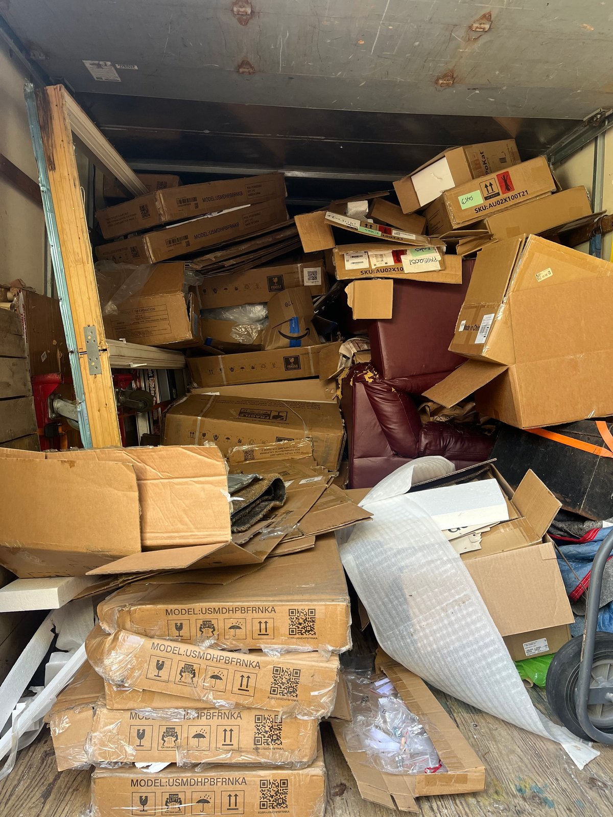 Cluttered storage unit or garage filled with stacked cardboard boxes, wooden furniture, and miscellaneous items