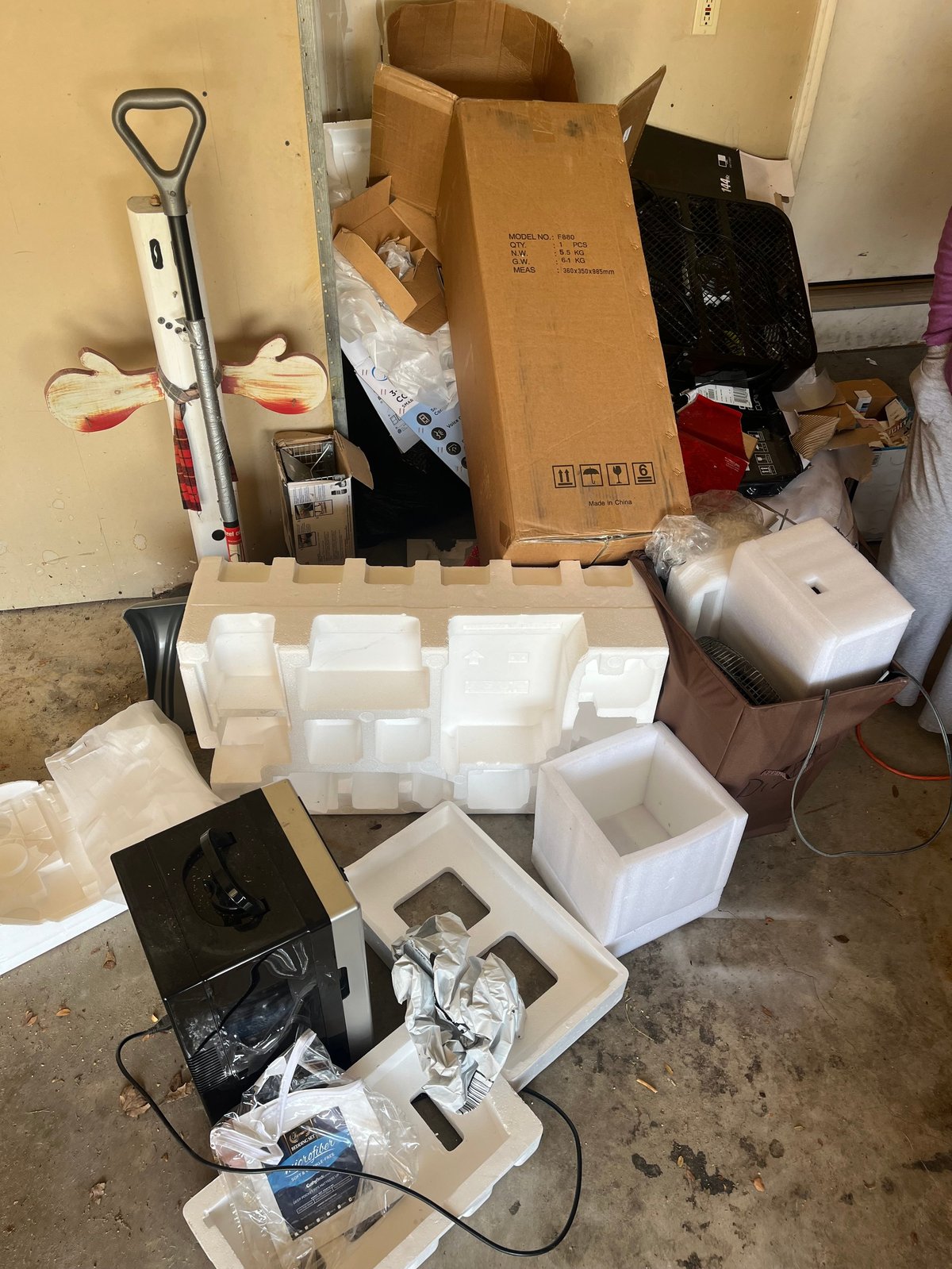 Cluttered garage or storage room with cardboard boxes, white foam containers, electronics, and a shovel against a beige wall