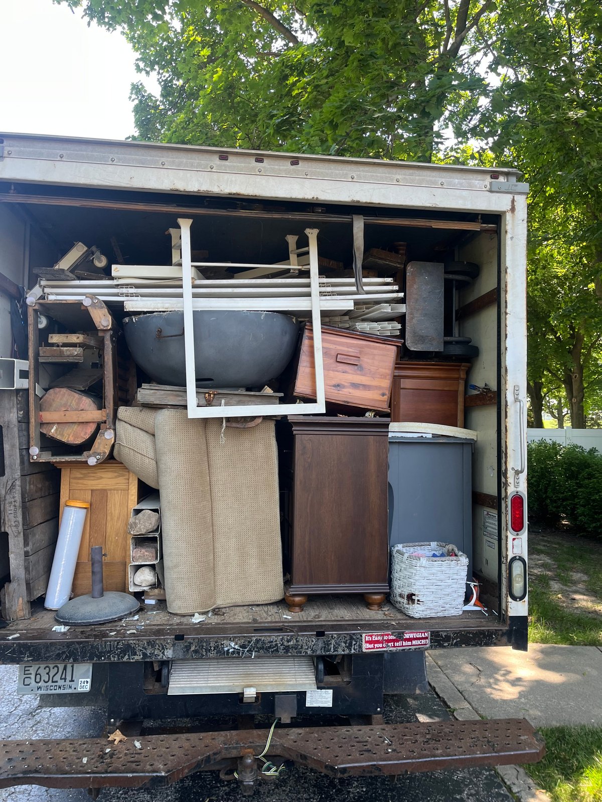 Open moving truck filled with various household furniture and belongings, parked on a residential street with green trees
